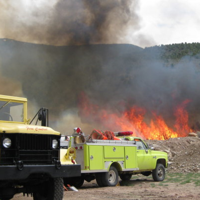 Yellow wildfire truck beside burning brush with thick smoke and flames behind it