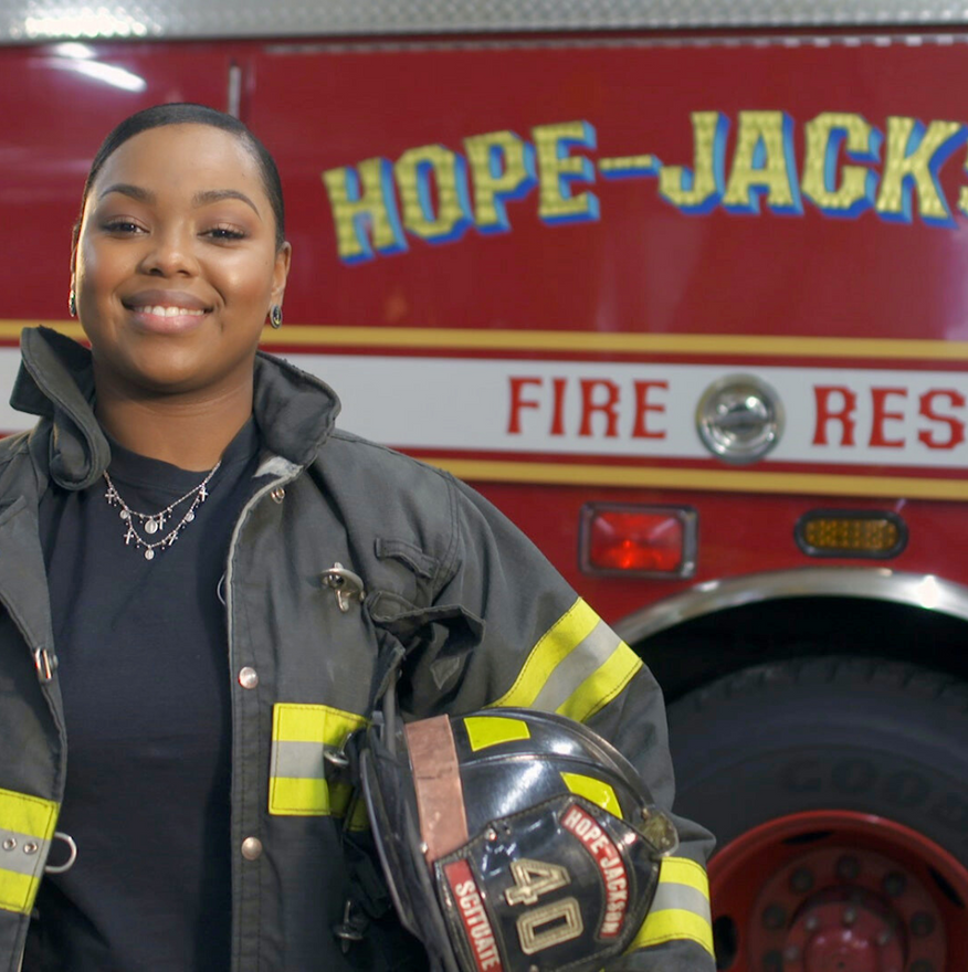 Smiling firefighter in black jacket holding a helmet beside a red fire rescue truck