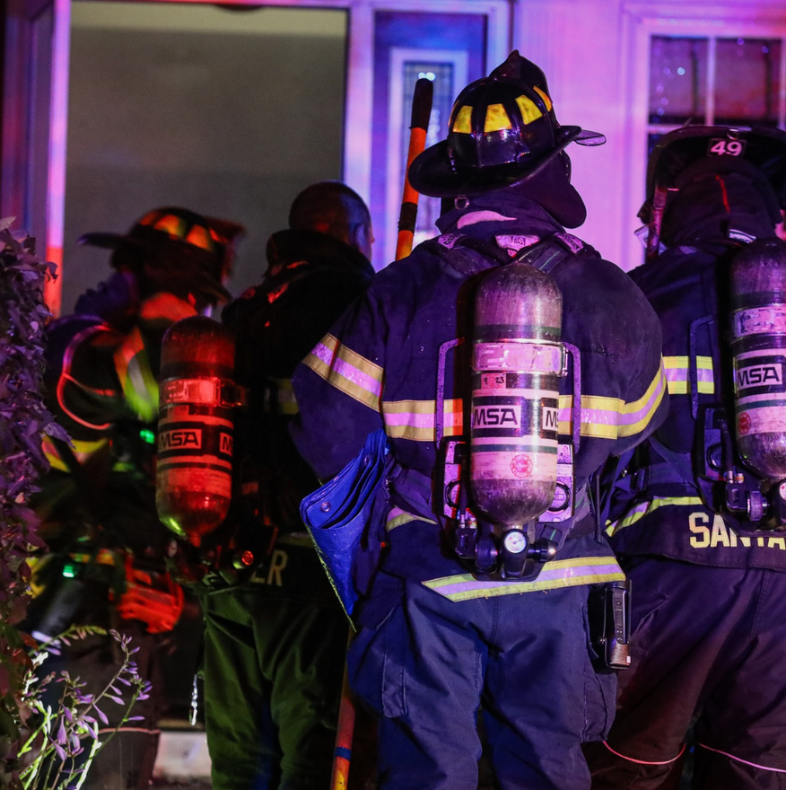 Firefighters in turnout gear responding at a smoke-filled building doorway at night.
