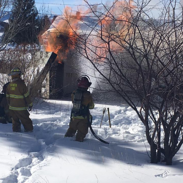Firefighters battle a house fire in a snowy yard, with orange flames and smoke rising behind bare trees