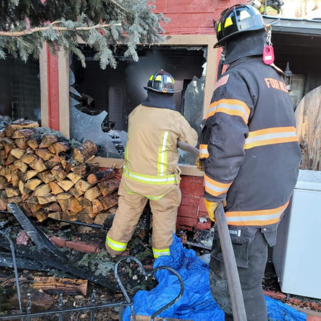 Firefighters inspect a charred building entrance beside stacked firewood and a blue tarp.