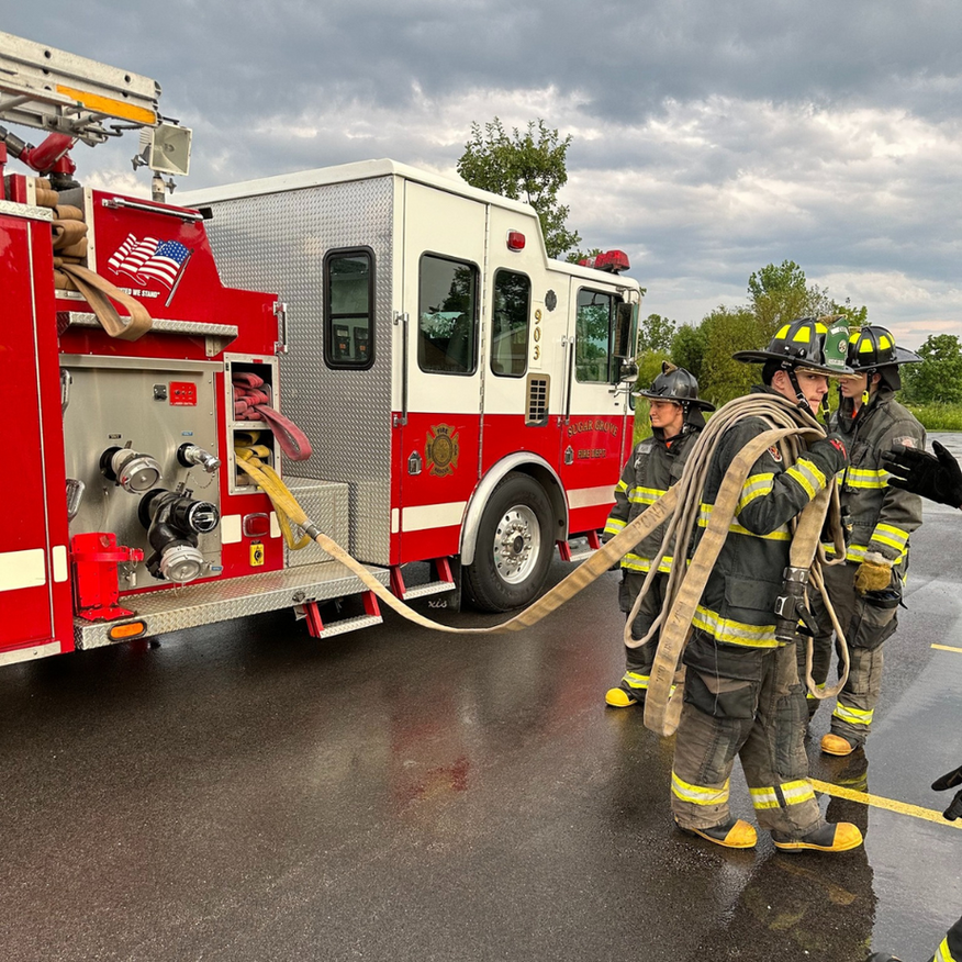 Firefighters in yellow gear work beside a red fire truck on a wet road under cloudy skies.