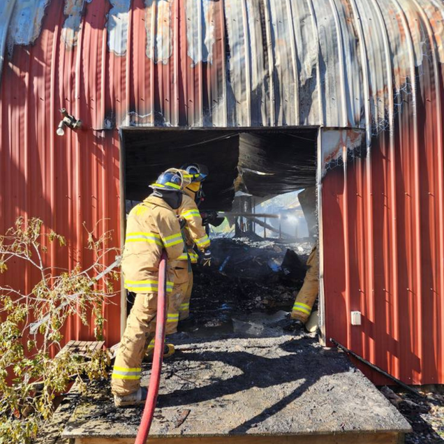 Firefighters in yellow gear work at a charred doorway of a red metal building