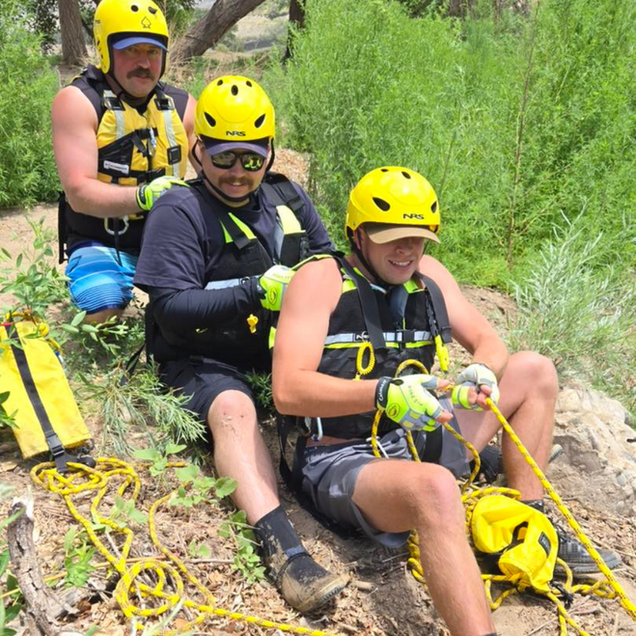 Three people in yellow helmets and harnesses sit on a rock, preparing climbing ropes outdoors.