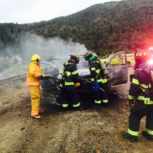 Firefighters in yellow gear extinguish a smoking car on a roadside with emergency lights flashing.