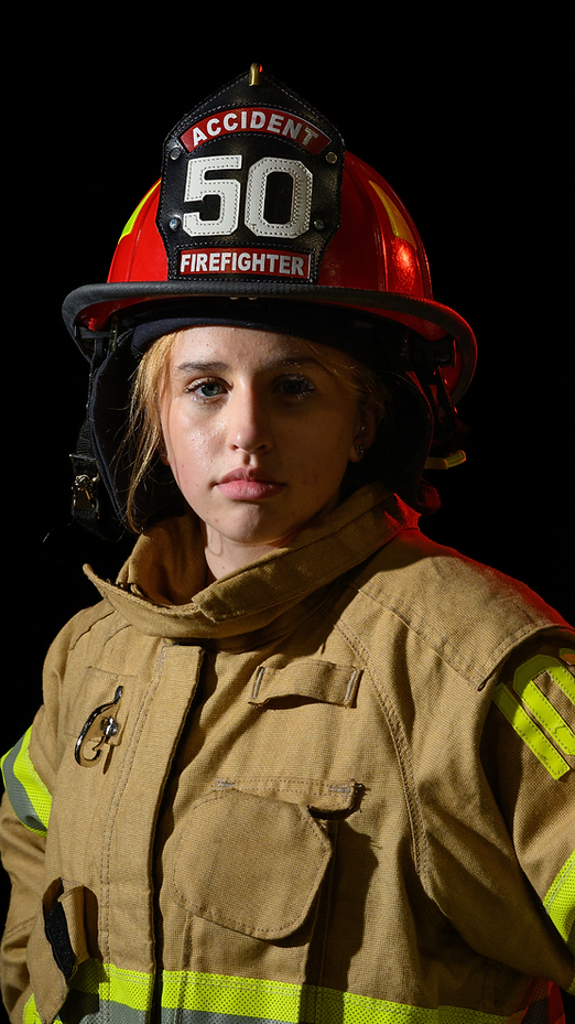 Firefighter in tan turnout gear and red helmet against a black background, facing forward.