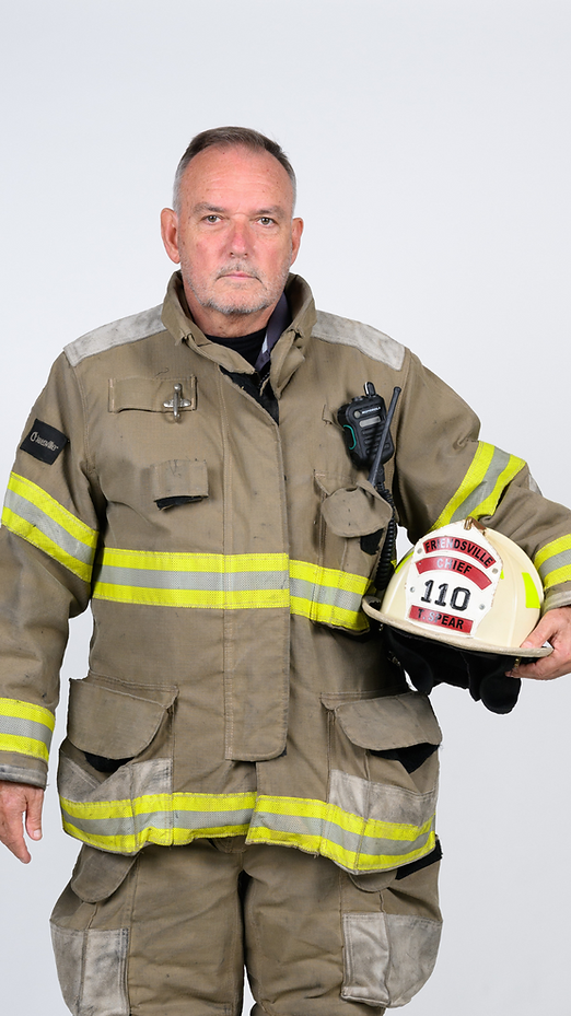 Firefighter in tan turnout gear holding a white helmet against a plain background