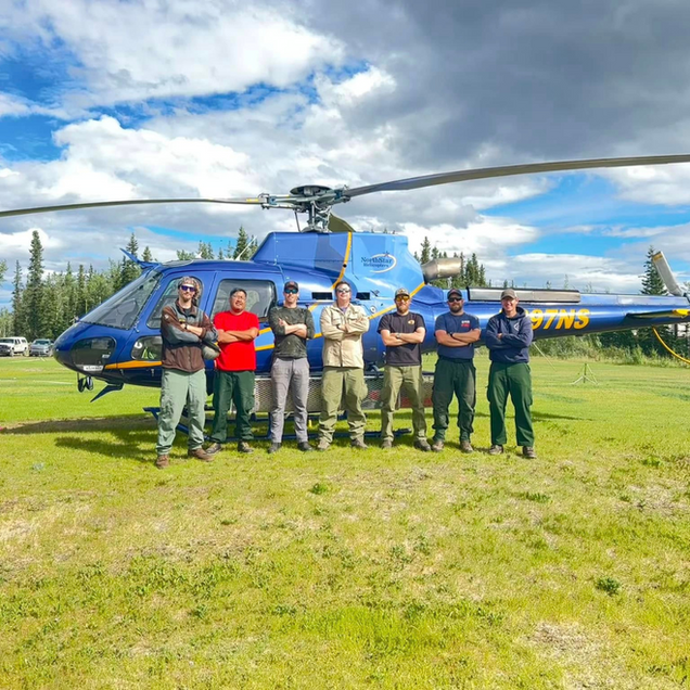 Seven people standing in front of a blue helicopter on a grassy field under a cloudy sky