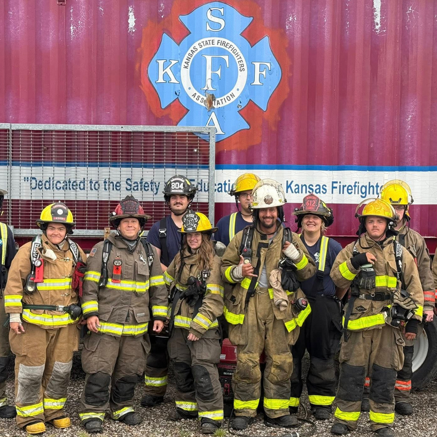 Group of firefighters in gear posing in front of a red building with a logo.
