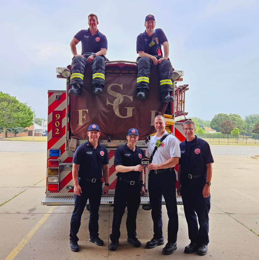 Firefighters posing in front of a red fire truck at a station, two seated on top and four standing below