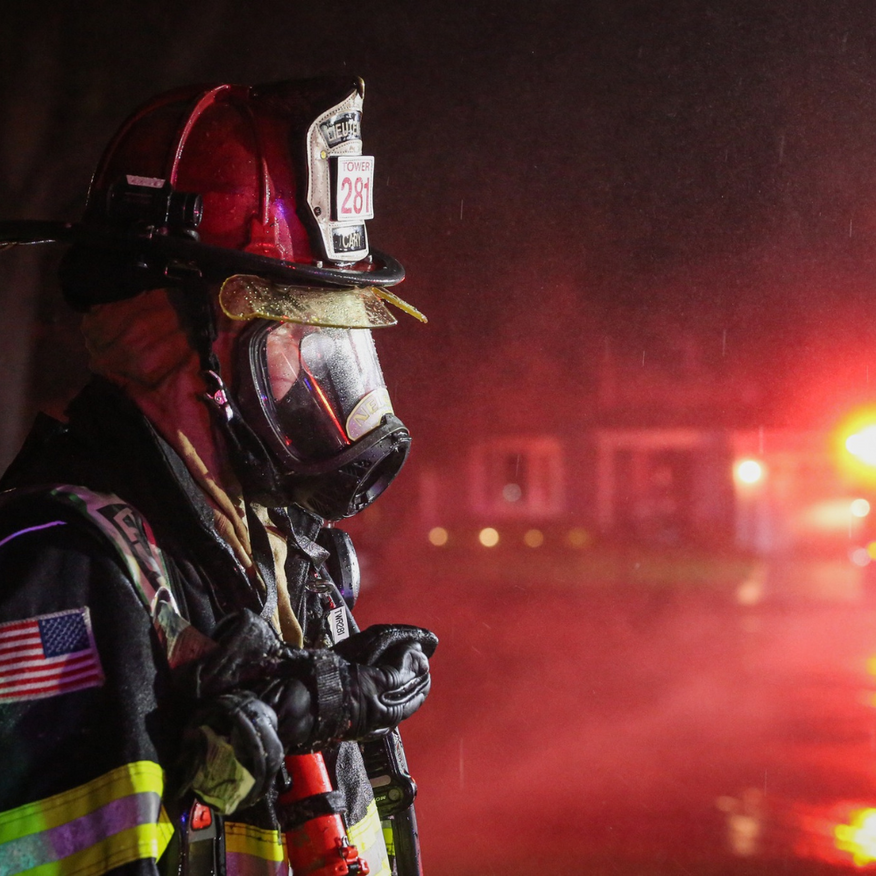 Firefighter in helmet and mask stands in red-lit smoke at a nighttime emergency scene.