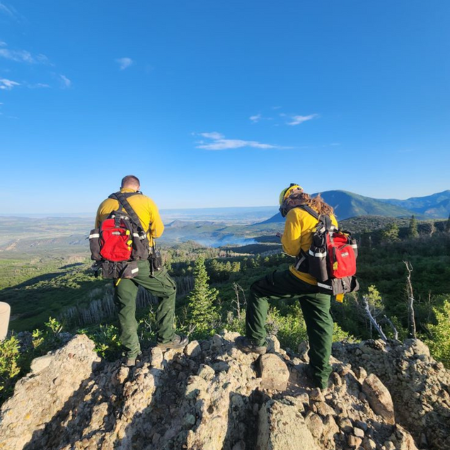 Two hikers with red packs stand on a rocky peak overlooking mountains and a valley under a blue sky