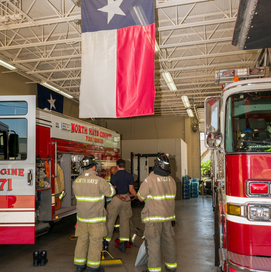Firefighters in tan gear walk through a fire station between red fire trucks under a Texas flag.