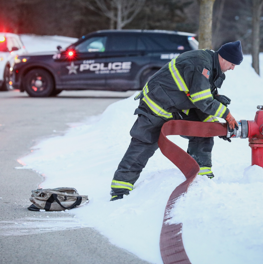 Firefighter in dark uniform operating a red hydrant beside a snowy road, police car in background