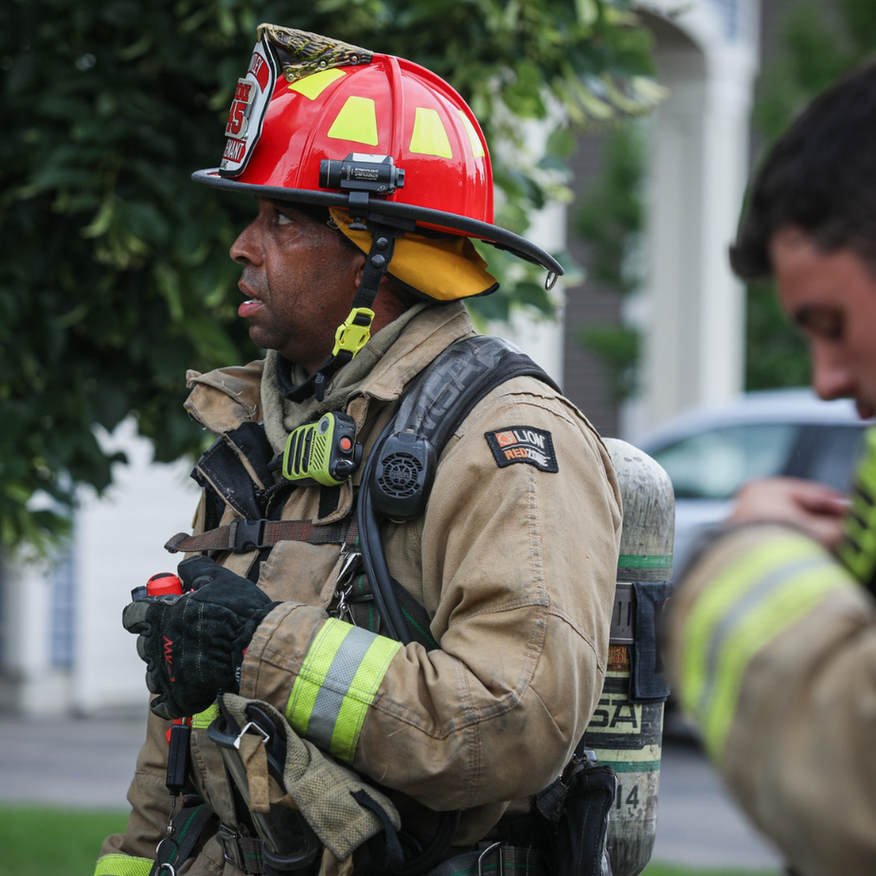 Firefighter in tan gear and red helmet standing outdoors, speaking on a street with a blurred figure nearby