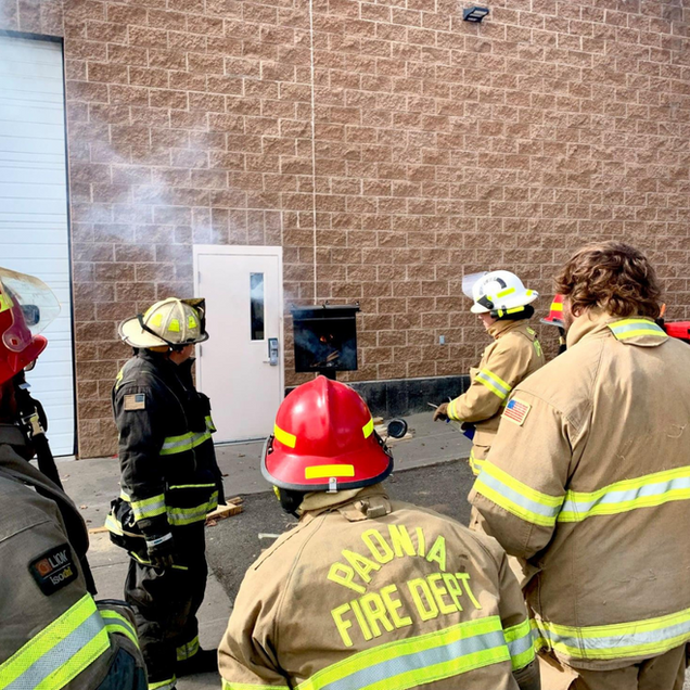 Firefighters in tan and black gear gather outside a brick building with smoke drifting from a door.