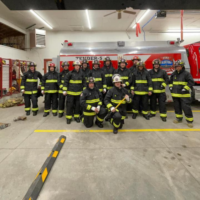Group of firefighters in black and yellow gear posing in a fire station garage with a fire truck behind them