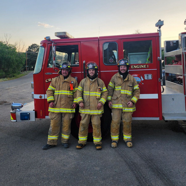 Three firefighters in yellow gear standing beside a red fire engine at dusk