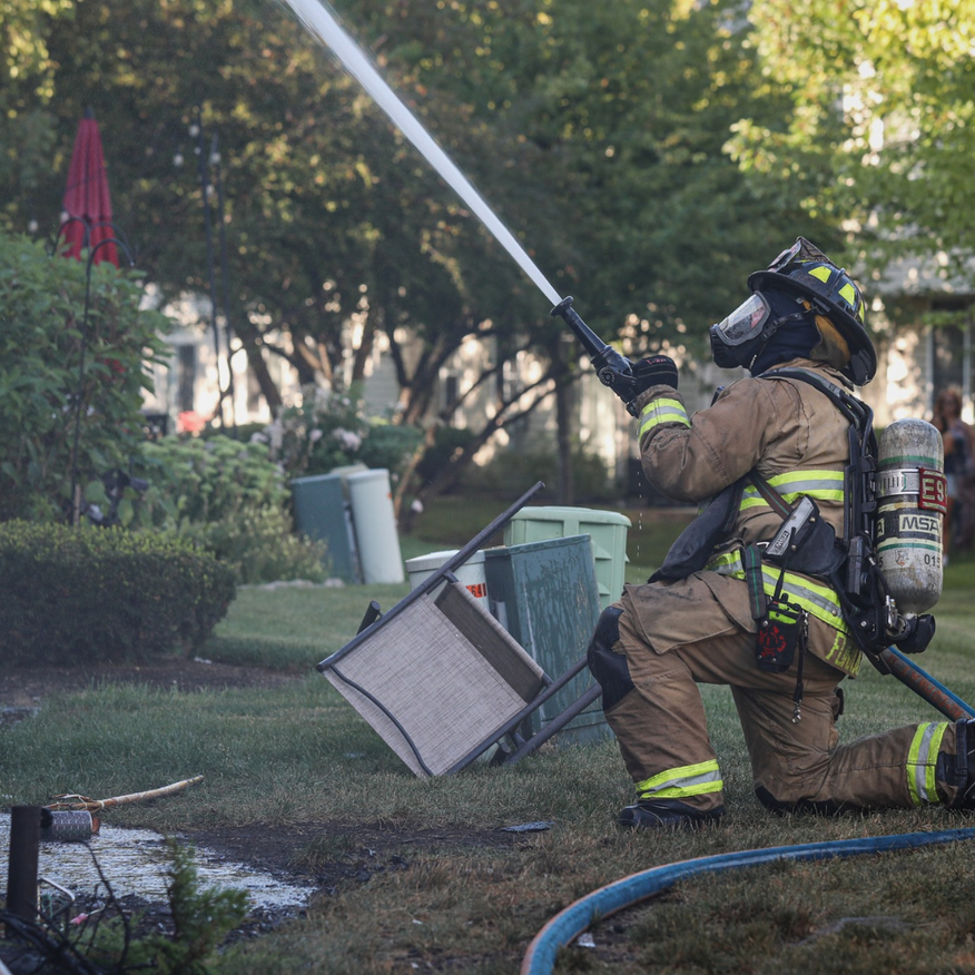 Firefighter kneeling and spraying water on a small grass fire near a toppled sign in a park
