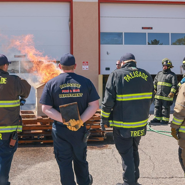 Firefighters in black gear stand outside a building, watching a controlled fire burn on stacked pallets.