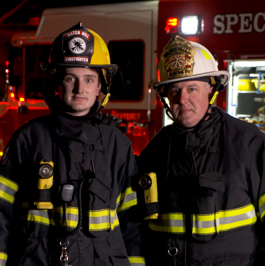 Two firefighters in black gear and helmets standing in front of a red fire truck at night