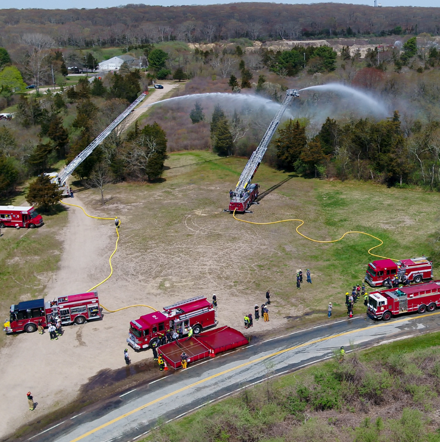 Firefighters spray water from two ladder trucks at a grassy field fire near a road and trees.