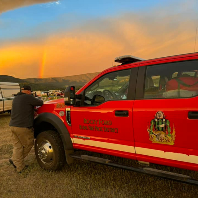 Red fire rescue truck with a person beside it at sunset on a grassy roadside