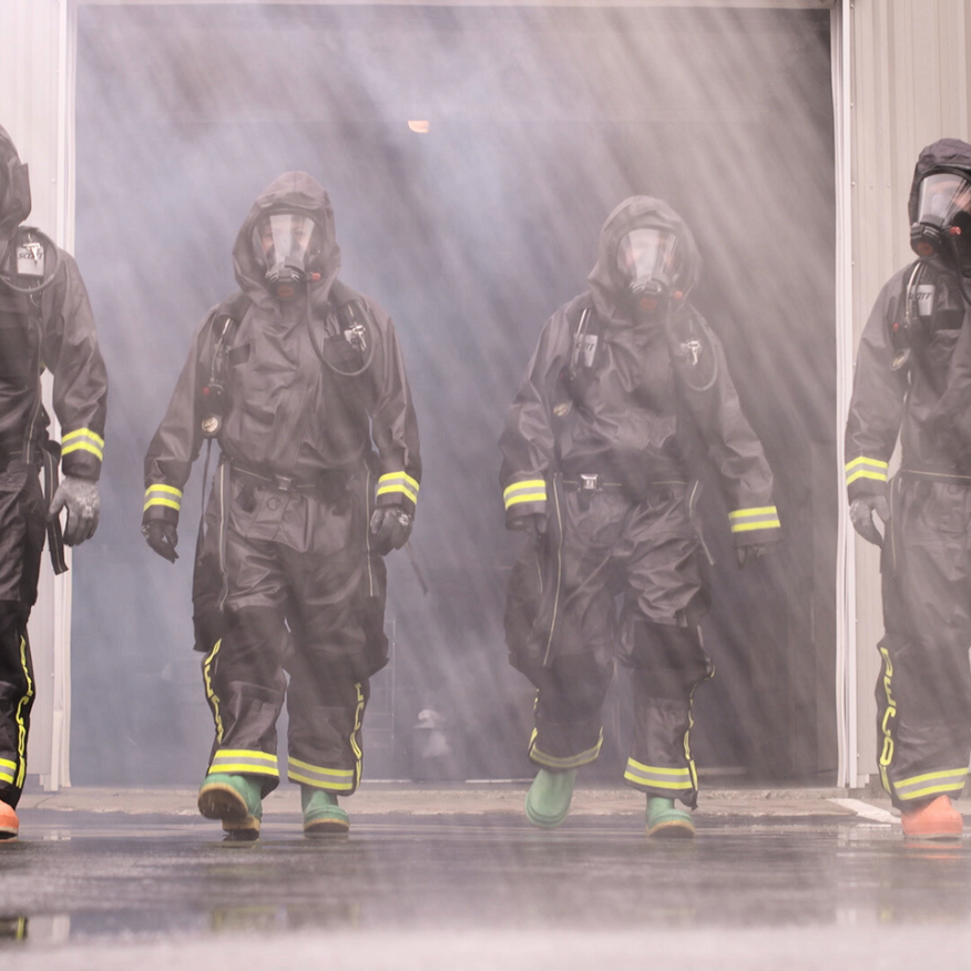 Firefighters in black gear emerge through heavy water spray from a smoky doorway.
