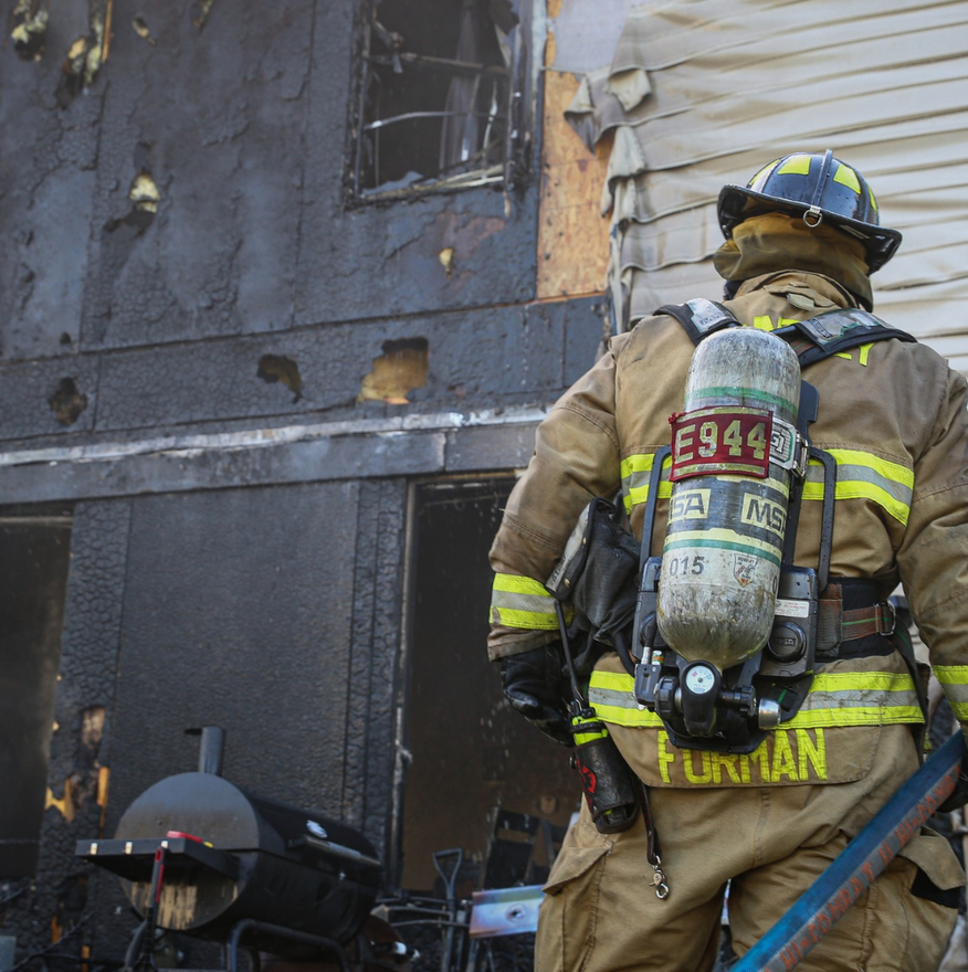 Firefighter in tan gear stands beside a smoke-damaged building, facing a charred doorway.