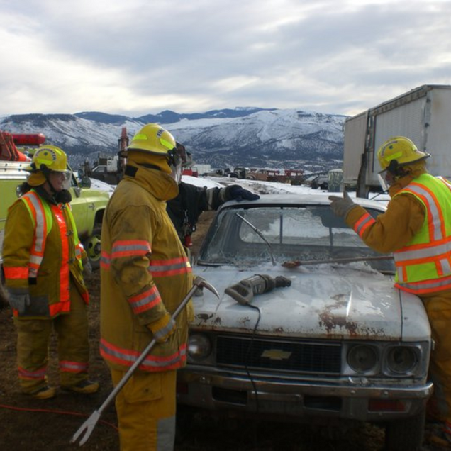 Firefighters in yellow gear inspect a burned car at an accident scene with snowy mountains behind them