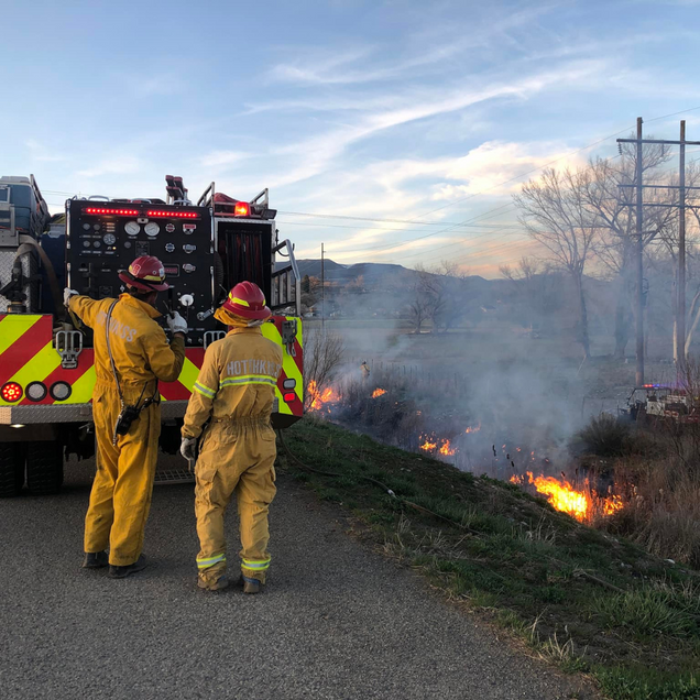 Two firefighters in yellow gear watch a roadside grass fire beside a fire truck at dusk.