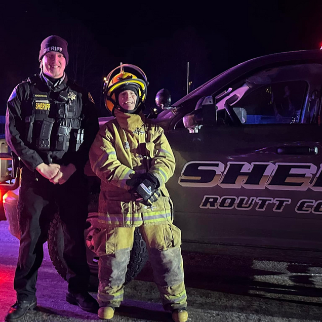 Two emergency responders in reflective gear stand beside a marked SUV at night under purple lighting