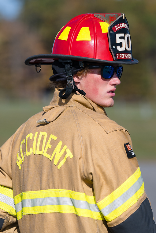 Firefighter in red helmet and tan jacket with “INCIDENT” on the back, wearing sunglasses, in profile.
