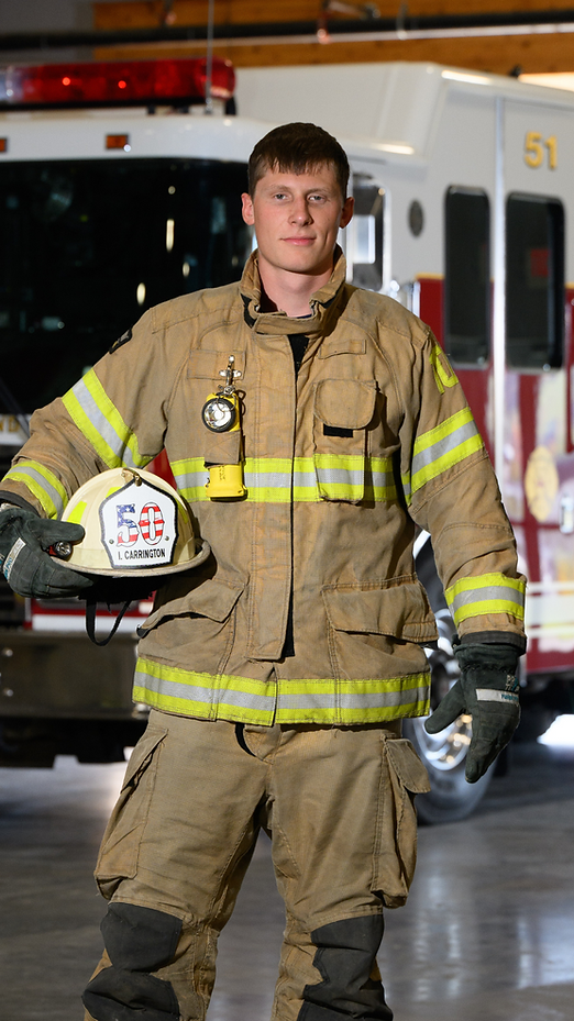 Firefighter in tan turnout gear holding a helmet, standing in front of a fire truck.