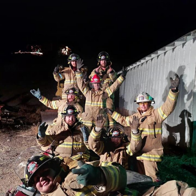 Firefighters in tan gear pose at night beside a white wall, flashing peace signs and thumbs-up.