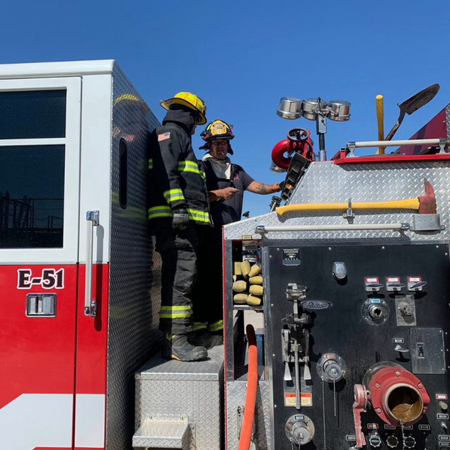 Firefighters in yellow helmets stand on a red fire truck, preparing equipment under a clear blue sky.