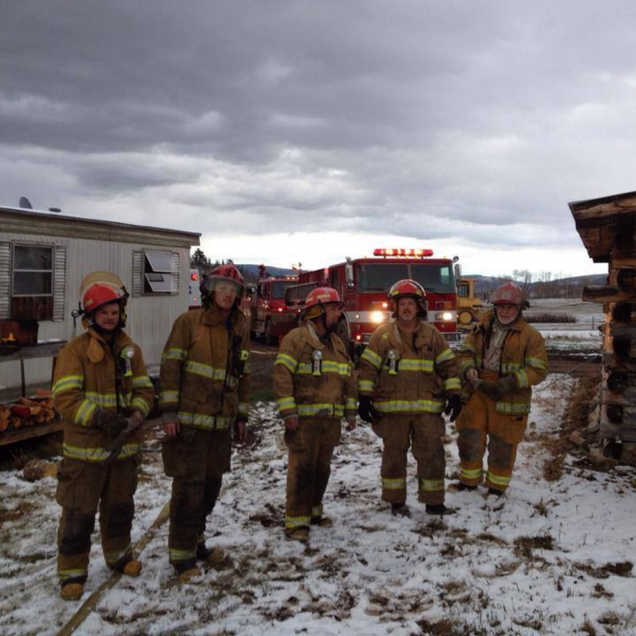 Five firefighters in yellow gear stand in a snowy yard beside fire trucks and a house.
