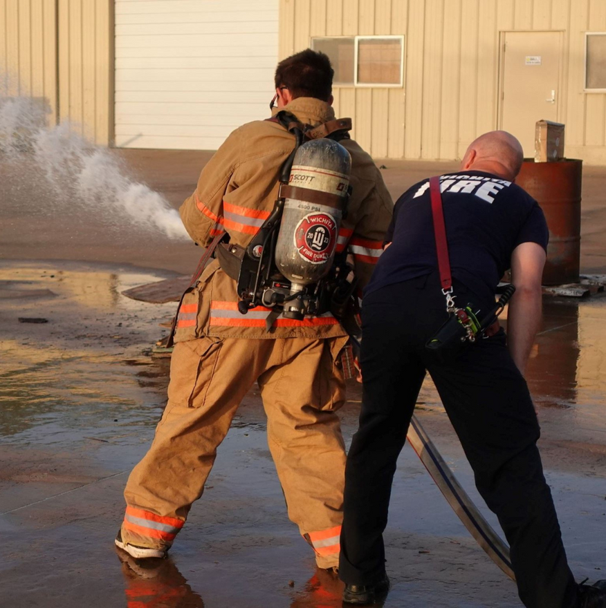 Two firefighters training with a water hose outdoors; one sprays water, another steadies the hose.