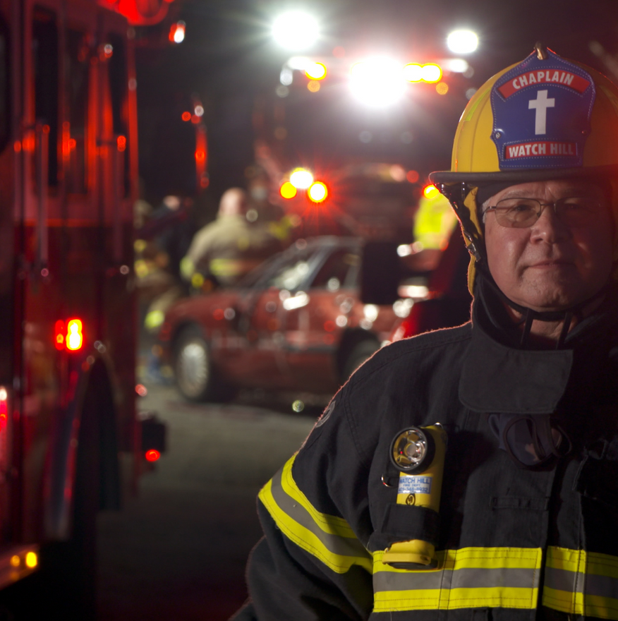 Firefighter in reflective gear stands beside an emergency vehicle at night with bright flashing lights.