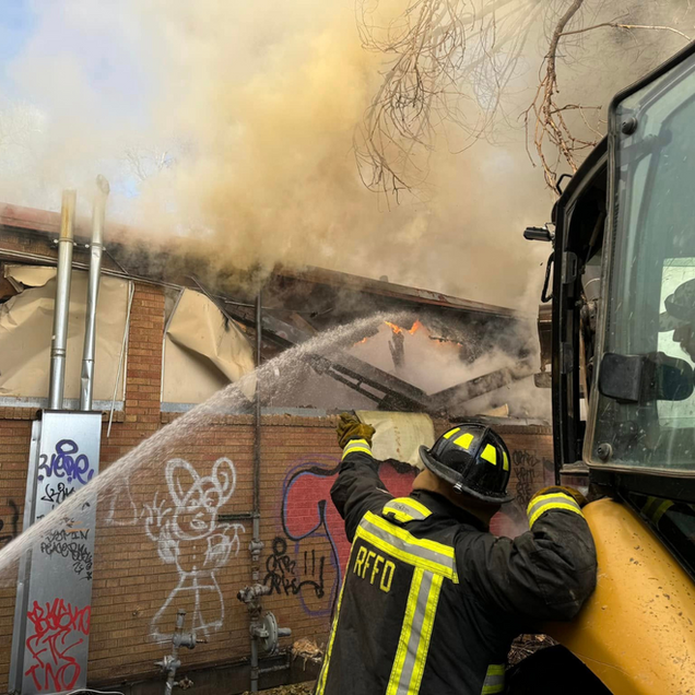 Firefighters spray water at a burning building beside a yellow truck, with heavy smoke billowing overhead.