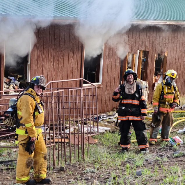 Firefighters in yellow and black gear stand outside a smoke-filled wooden house after a fire.