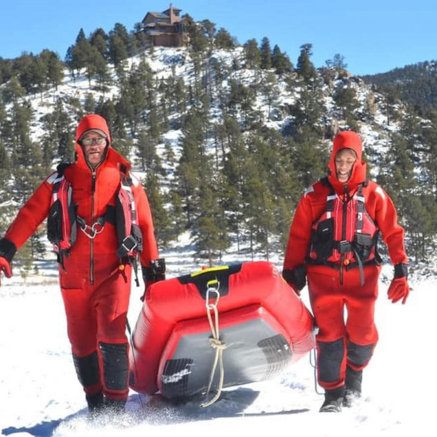 Two people in red cold-weather gear pull an inflatable sled across snowy terrain with a cabin in the background