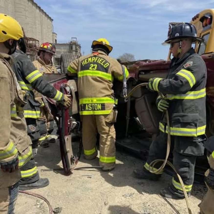 Firefighters in yellow helmets work together to extricate someone from a crushed car at a roadside scene.