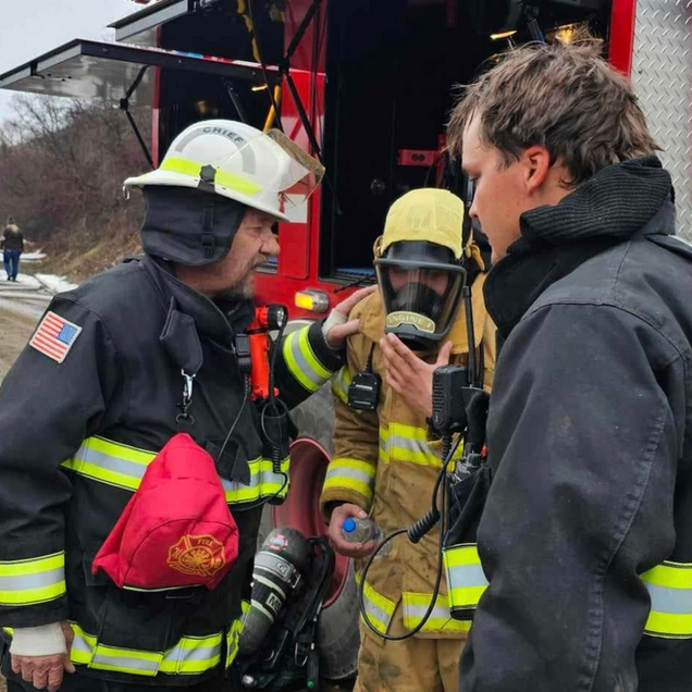 Firefighters in gear conferring beside a red truck, one holding a radio while a man stands nearby.