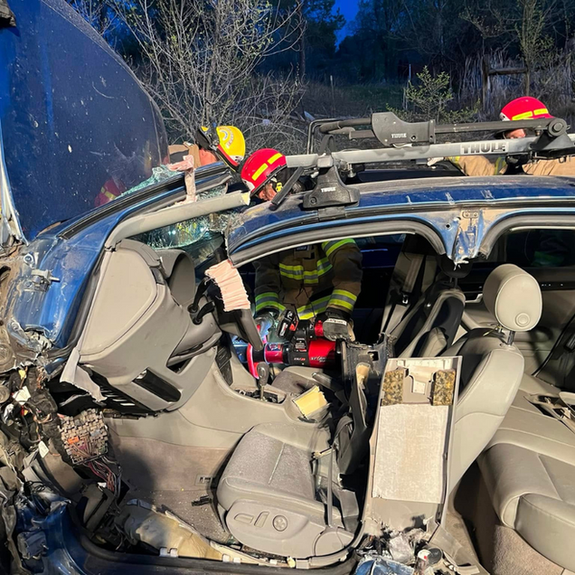 Severely damaged car with roof crushed, firefighters in helmets, and the interior exposed at a crash scene
