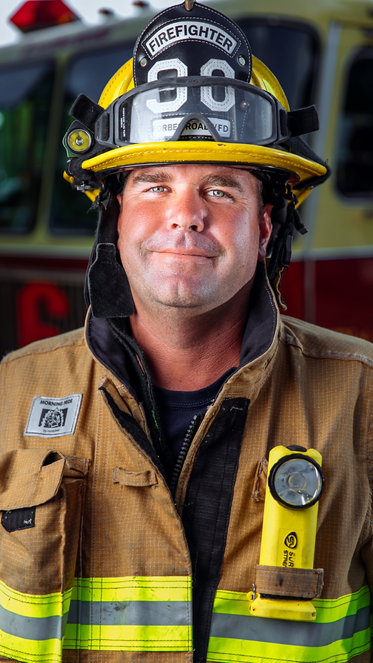 Firefighter in tan turnout gear and black helmet, standing in front of a fire truck, slight smile.