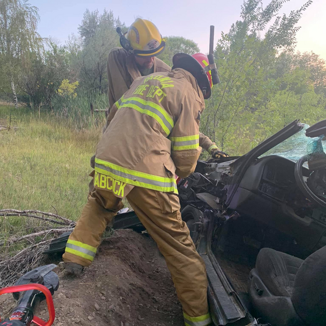 Firefighters in tan gear working to free a car from a ditch beside brush and trees