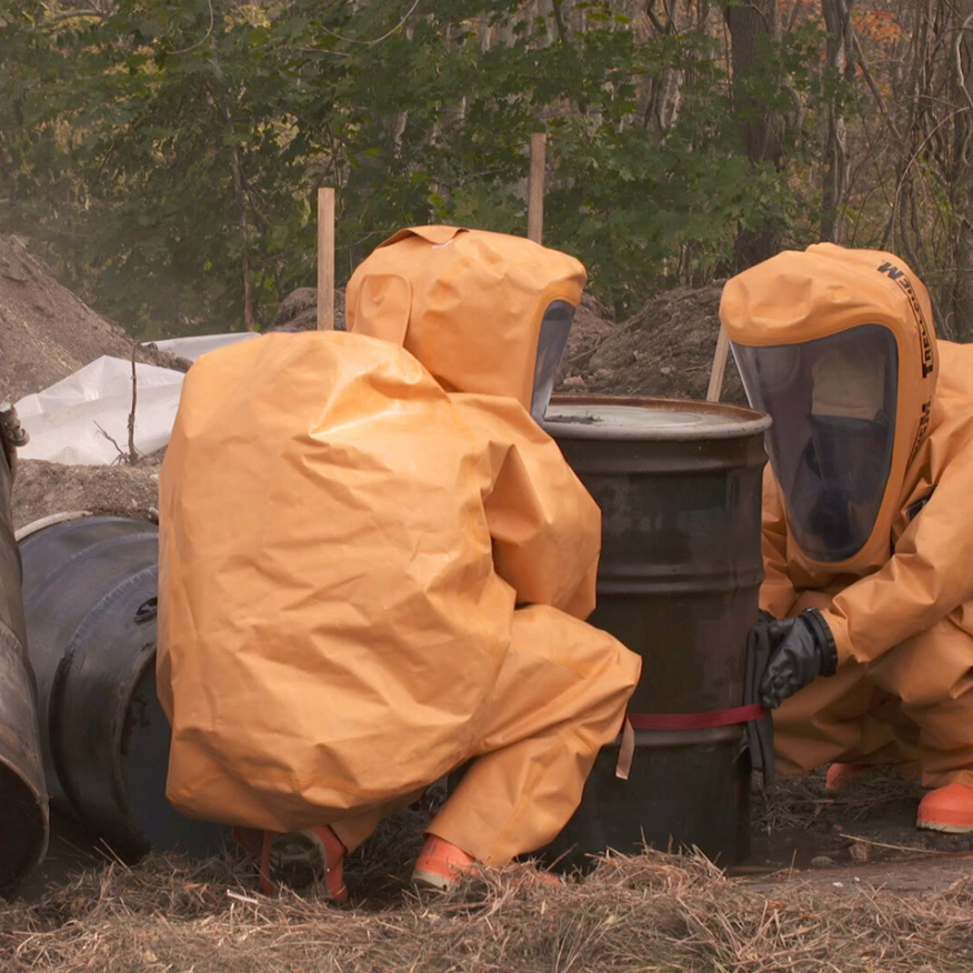 People in orange hazmat suits around a black barrel outdoors, near fallen trees and brush.