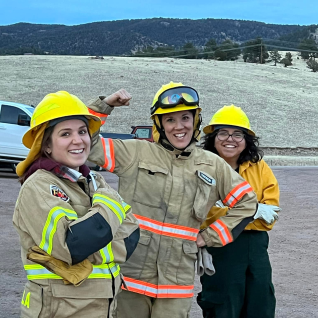 Three people in safety gear smiling outdoors at a rocky work site, wearing hard hats and reflective jackets