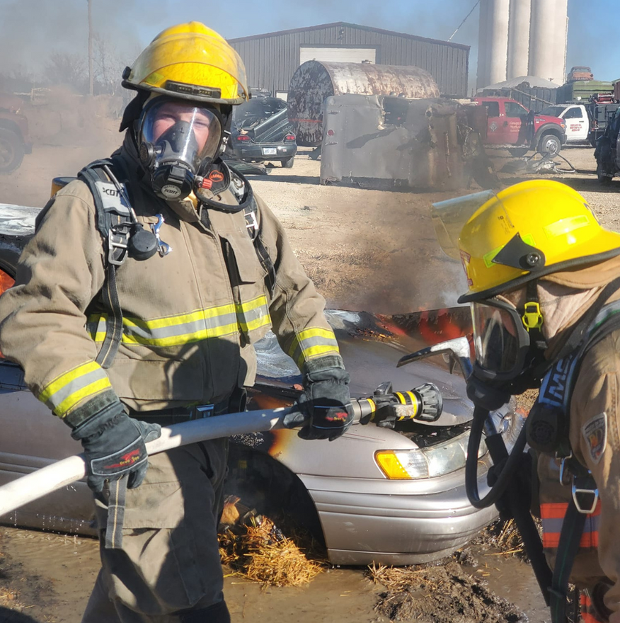 Two firefighters in gear extinguishing a car fire with water; outdoor setting.